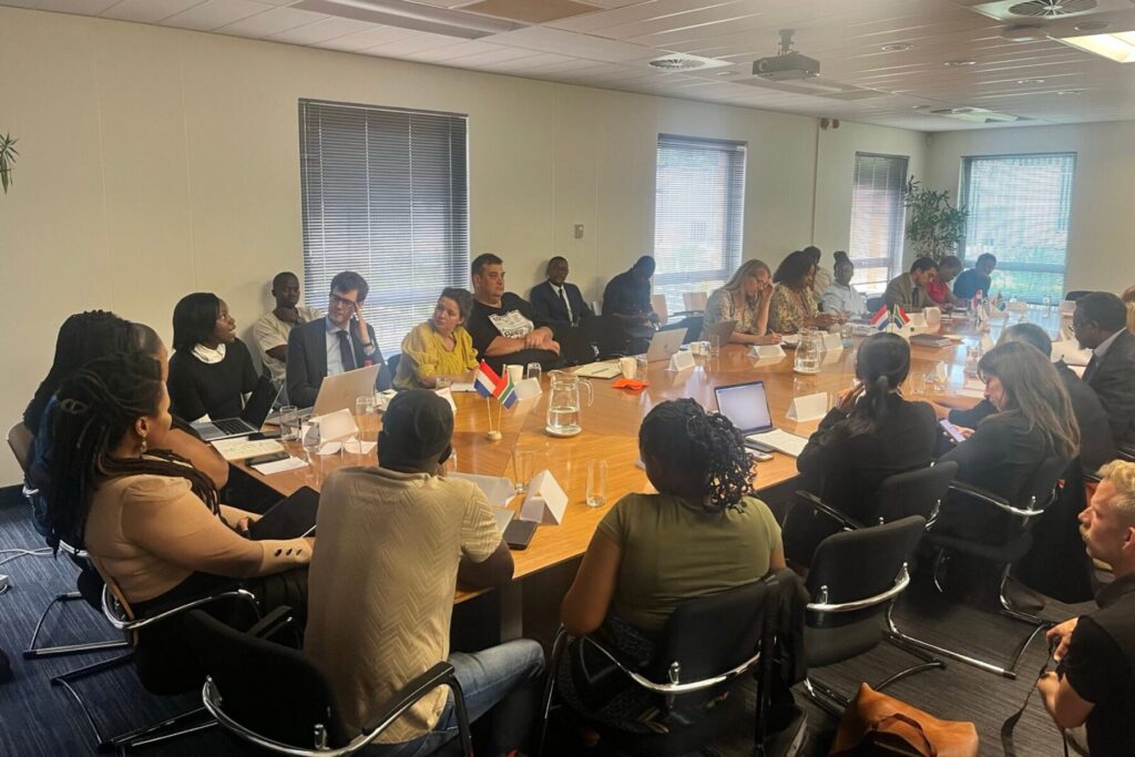 A meeting around a large wooden conference table with participants including Special Rapporteur Mary Lawlor and human rights defenders from South Africa, Eswatini, Kenya, Mozambique, Lesotho, and Zimbabwe, organised by the Dutch embassy in South Africa and the African Initiative of Women Human Rights Defenders. The room is well-lit, with windows on one side. Small table flags of South Africa and the Netherlands are placed on the table. Participants are engaged in discussion or taking notes, with notebooks, glasses of water, and papers scattered across the table.