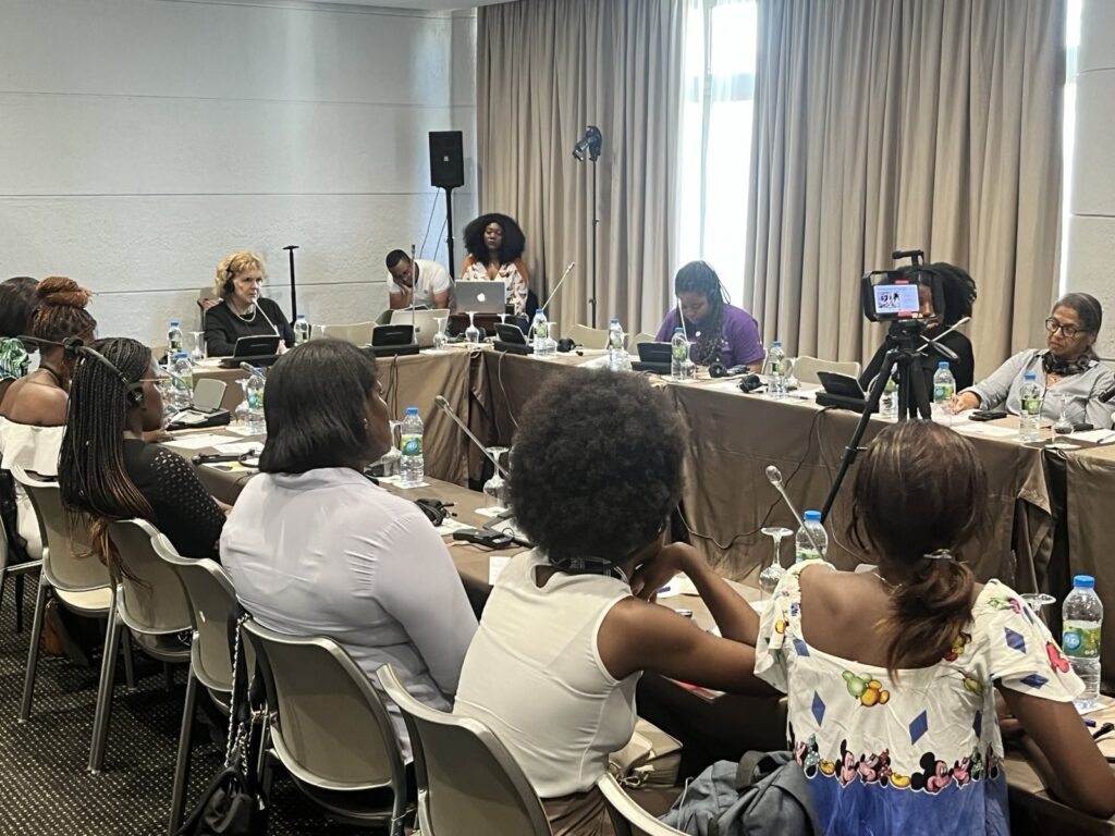 A meeting featuring the UN Special Rapporteur on Human Rights Defenders Mary Lawlor and Angolan women human rights defenders. The photo shows a diverse group of women seated around a rectangular table covered with brown tablecloths, engaged in discussion. Bottled water, microphones, and notepads are visible on the table. A camera is positioned in the foreground for recording. A woman stands at the back near a laptop, while another participant takes notes. Curtains partially cover a window in the background, allowing natural light to enter the room.