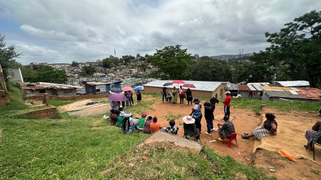 A group of people gathered outdoors on a grassy slope in Cato Crest, South Africa, for a meeting between UN Special Rapporteur on Human Rights Defenders Mary Lawlor and members of the Abahlali baseMjondolo movement. Some are seated on chairs or the ground, while others stand holding umbrellas. Behind them are informal housing structures made of corrugated metal and wood, with a dense settlement visible in the background.