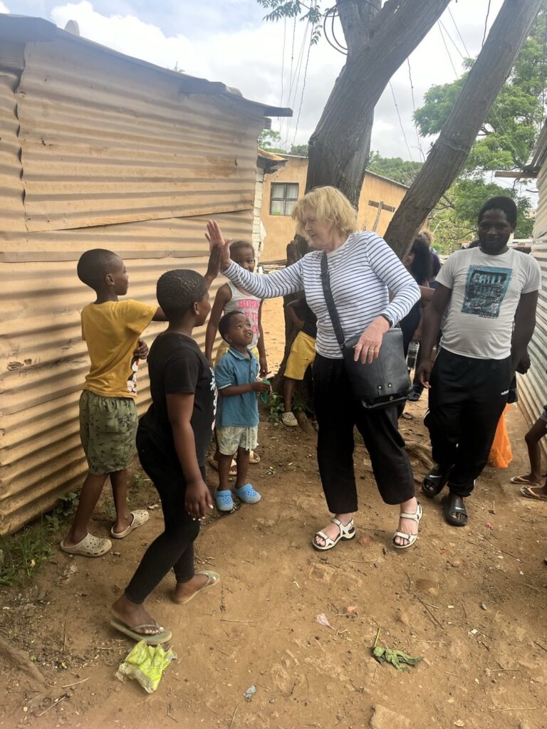 UN Special Rapporteur Mary Lawlor interacting with children during a visit to the Cato Crest branch of Abahlali baseMjondolo community, near Durban in South Africa. She is smiling and giving a high-five to one of the children, who are gathered around her near a corrugated metal shack. 