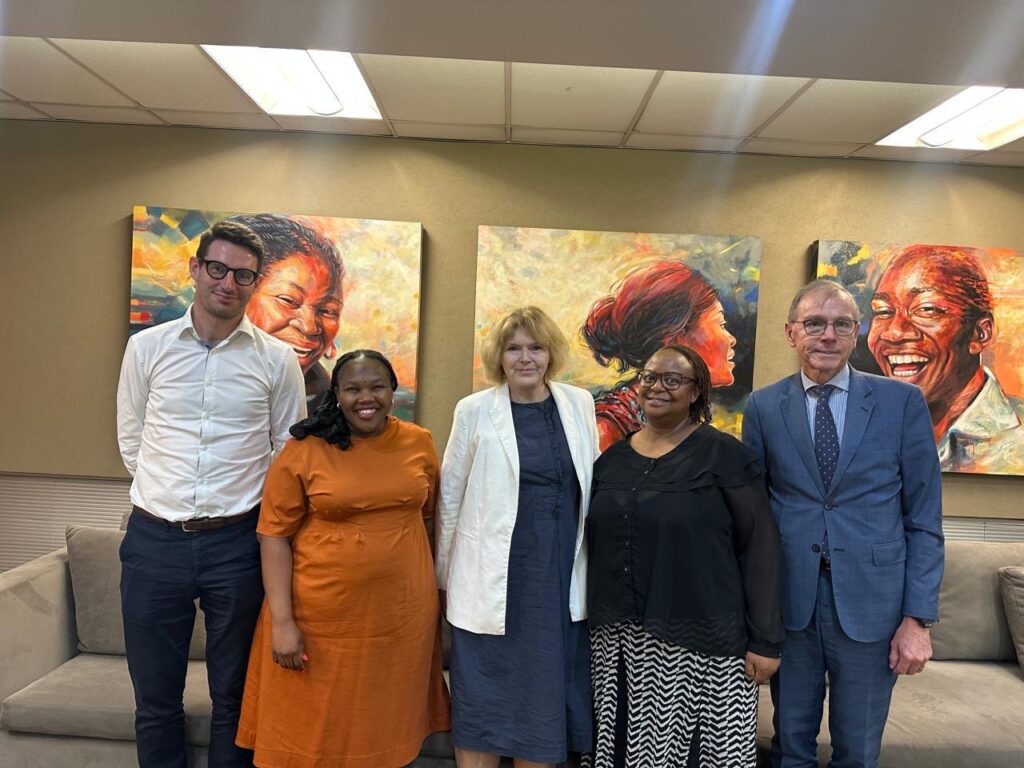 UN Special Rapporteur Mary Lawlor posing for a group photo after a meeting with three officials from South Africa's Department of International Relations and Cooperation (DIRCO) and a member of her team. The group is indoors, with vibrant, colourful paintings of smiling faces on the wall behind them.