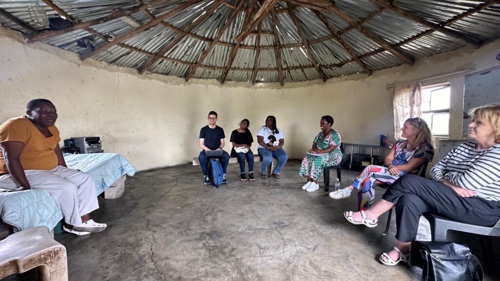 A meeting inside a circular thatched-roof building with concrete walls, involving UN Special Rapporteur on human rights defenders Mary Lawlor and community members affected by the Tendele coal mine in KwaZulu-Natal. The group is seated in a circle on plastic chairs, engaging in conversation. The room has simple furnishings including a table, a sound system, and a curtain-covered window letting in natural light.