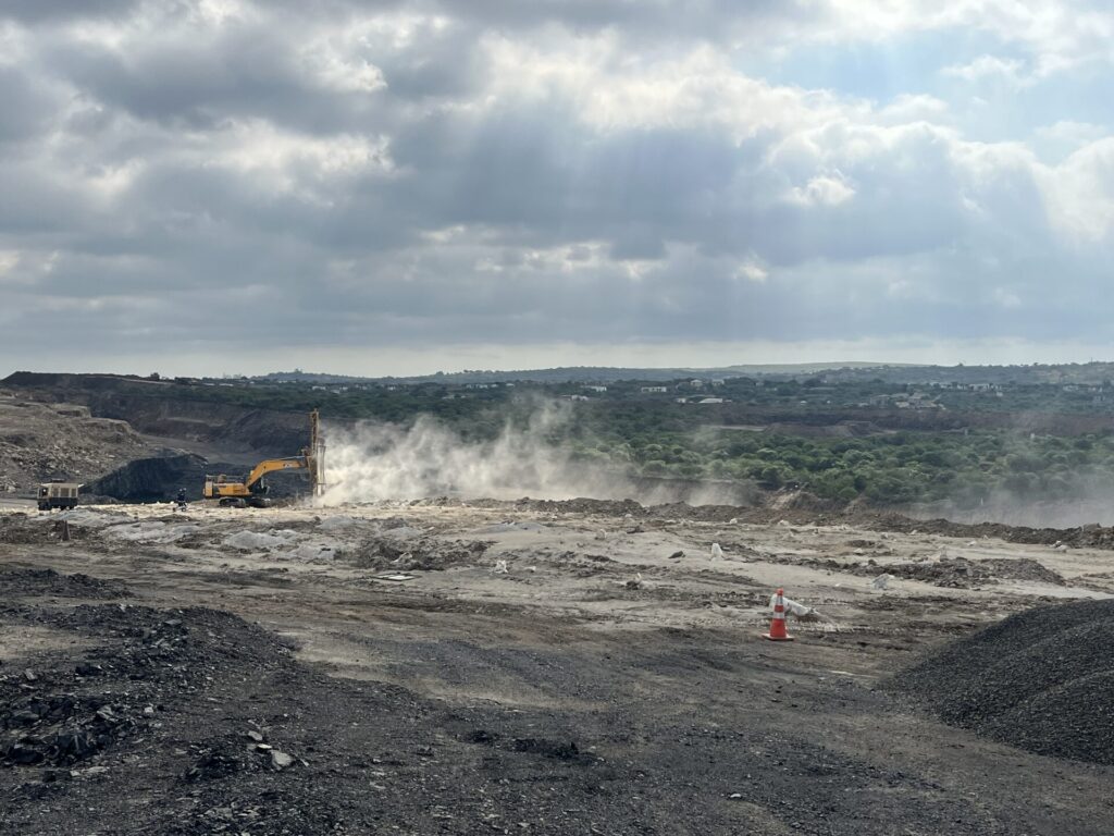 A photo taken at the Tendele coal mine site in KwaZulu-Natal. Mining operations are ongoing, creating clouds of dust. The ground is dry and rocky, and there are surrounding hills stretching into the distance.