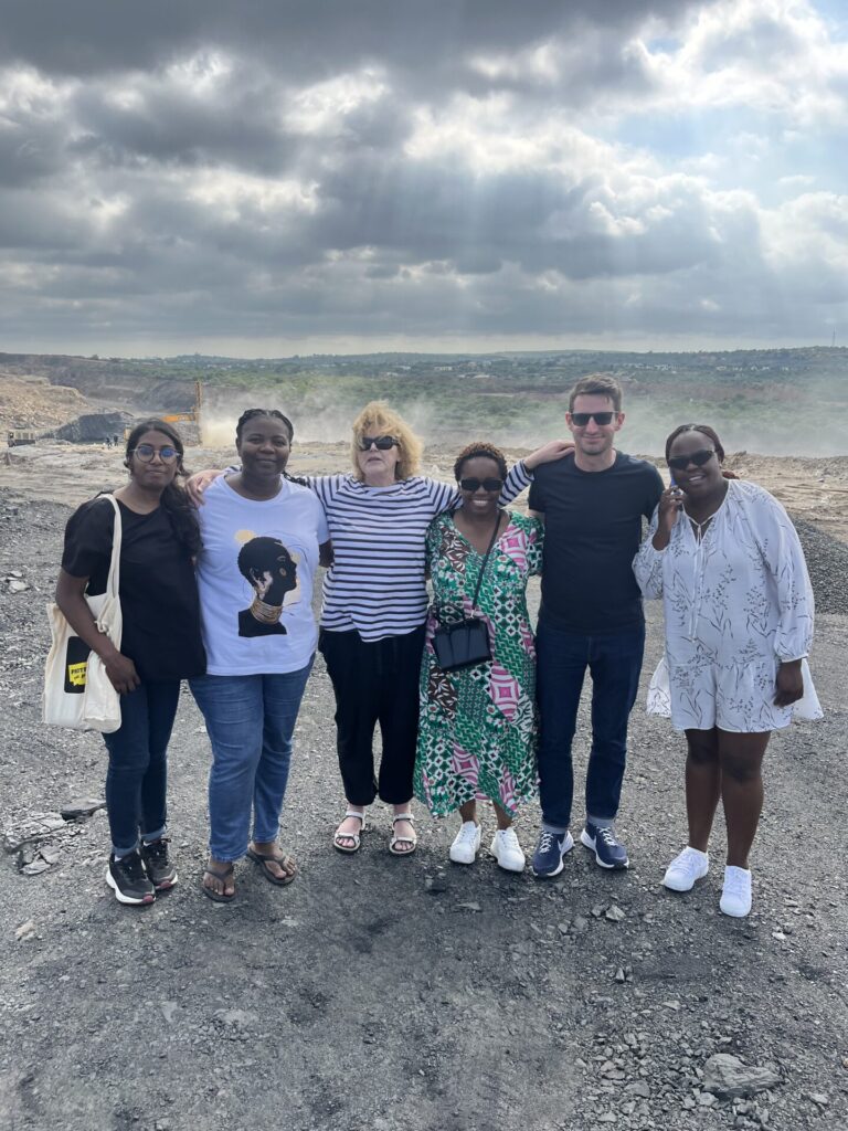 A group photo taken at the Tendele coal mine site in KwaZulu-Natal. Six individuals, including UN Special Rapporteur Mary Lawlor, stand closely together on a dusty, rocky surface, with a mining operation visible in the background under a cloudy sky. The group smiles warmly, with the landscape of the mine and surrounding hills stretching into the distance.