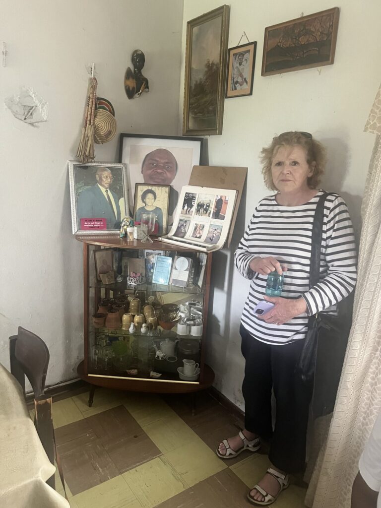 UN Special Rapporteur on human rights defenders Mary Lawlor is standing indoors near the corner of a house where framed photographs and various items are displayed on a table, in what looks like a shrine. She has a serious expression on her face.