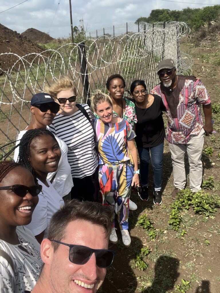 A group photo with nine individuals, including UN Special Rapporteur Mary Lawlor, standing closely together by a wired fence, on the other side of which is the Tendele coal mine site in KwaZulu-Natal.