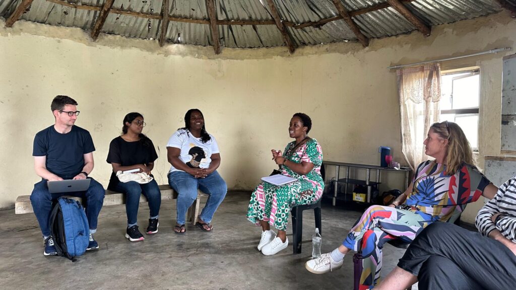 A meeting inside a circular thatched-roof building with concrete walls, involving UN Special Rapporteur on human rights defenders Mary Lawlor and community members affected by the Tendele coal mine in KwaZulu-Natal. The group is seated in a circle on plastic chairs, engaging in conversation. 