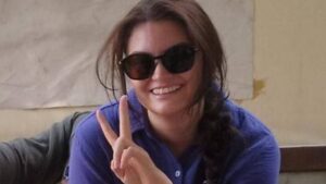 American-Turkish woman human rights defender Aysenur Ezgi Eygi, wearing sunglasses and a purple shirt, smiling and holding up a peace sign. She has long dark hair styled in a braid.