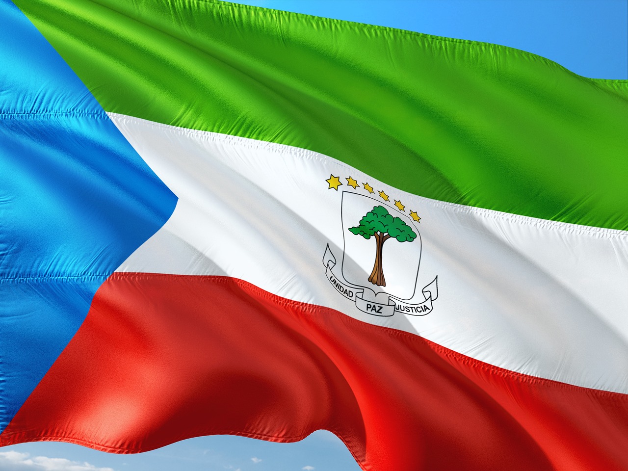 The flag of Equatorial Guinea, a horizontal tricolor of green, white and red with a blue isosceles triangle based on the hoist side and the National Coat of arms of Equatorial Guinea centered in the white band, flying in the wind.