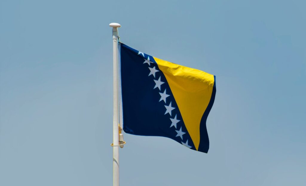 The flag of Bosnia and Herzegovina flying from the top of a pole against a blue sky background.