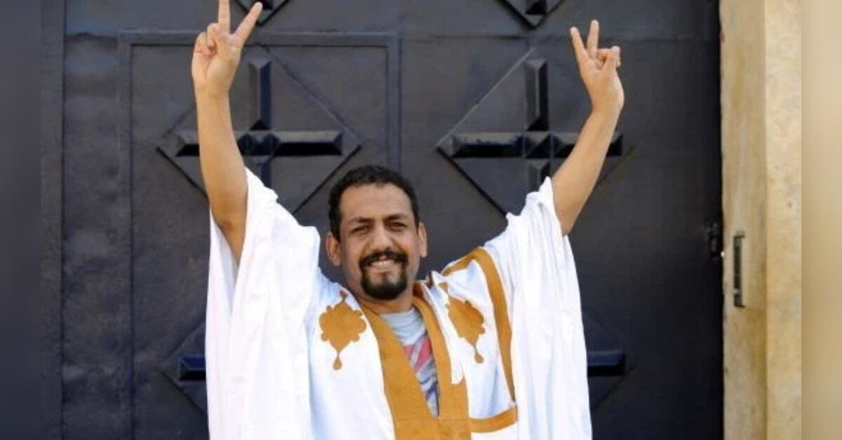 Western Sahara human rights defender Ali Salem Tamek stands in front of a dark door wearing a white and gold traditional Sahrawi attire. He smiles and raises both arms in a double peace sign gesture.