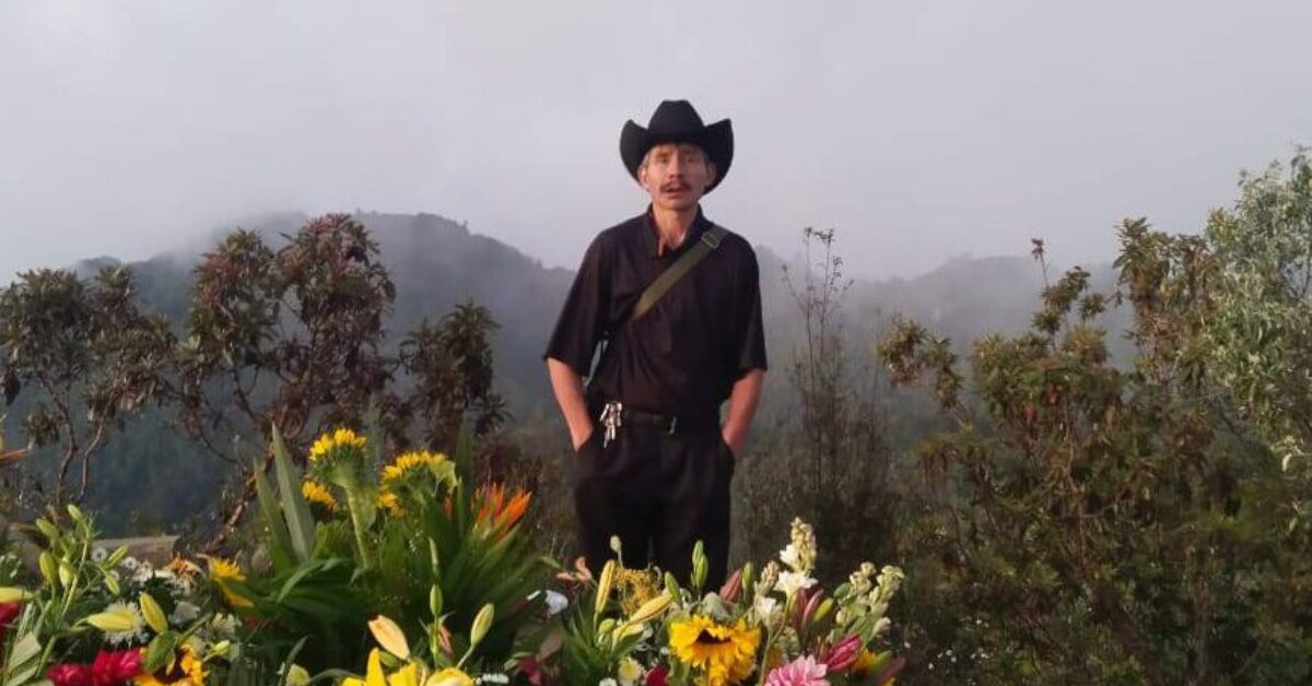 Mexican human rights defender Mario Gómez López stands outdoors in a misty mountainous landscape, wearing a black shirt and cowboy hat, with a strap across his shoulder and keys clipped to his belt. He is surrounded by vibrant flowers in the foreground, with trees and hills fading into the mist behind him.