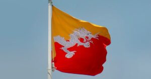 The flag of Bhutan flying from a pole against a bright blue sky.