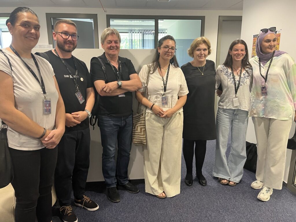 UN Special Rapporteur on human rights defenders Mary Lawlor stands amongst a group of six human rights defenders who work on the rights of migrants, refugees and asylum seekers in Bosnia and Herzegovina. The group is gathered in an office setting, smiling and wearing visitor badges.