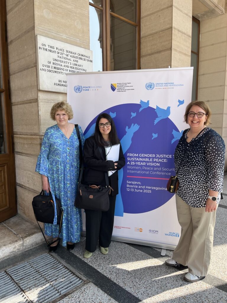 Mary Lawlor, UN Special Rapporteur on human rights defenders, stands with two members of her team in front of a banner for the international conference “From Gender Justice to Sustainable Peace: A 25-Year Vision,” held on 12-13 June 2025 in Sarajevo. They are smiling and standing outdoors beside the historic city hall building, with a commemorative plaque visible on the wall behind them.