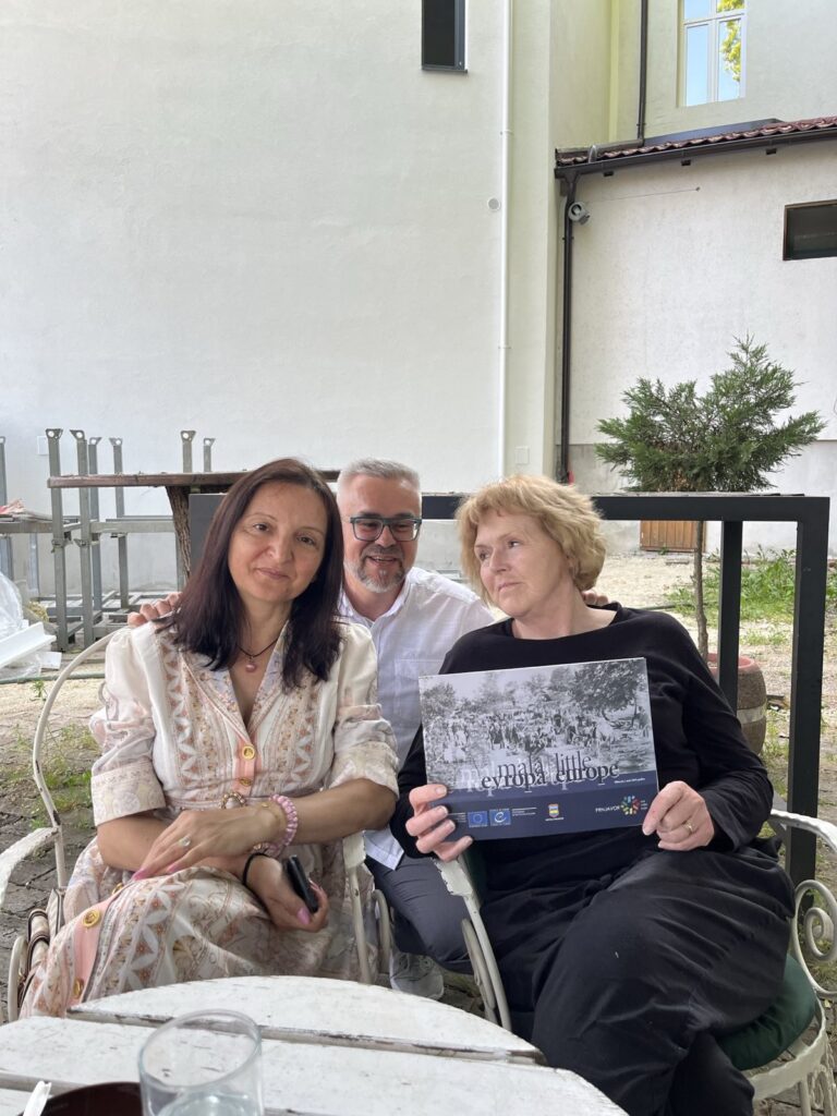 UN Special Rapporteur on human rights defenders Mary Lawlor sits with two human rights defenders from the Roma and Polish communities, who work to protect the rights of minorities. They are seated outdoors at a white table, smiling together. Mary holds a publication titled Mala Evropa / Little Europe, featuring historical images.