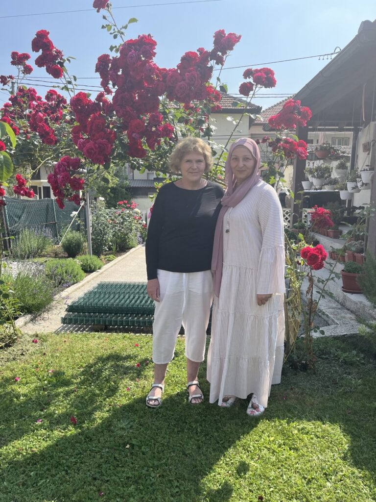 Mary Lawlor, UN Special Rapporteur on human rights defenders, stands with woman human rights defender Hajrija Čobo in a sunlit garden. They are surrounded by vibrant red rose bushes, with homes and hanging flower pots visible in the background. Both are smiling warmly on a clear, bright day.