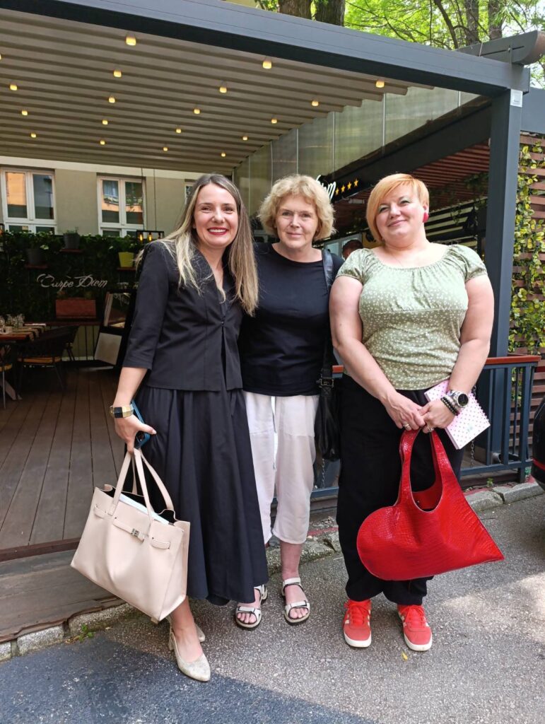 UN Special Rapporteur on human rights defenders Mary Lawlor stands with two women’s rights defenders in Zenica, during her official visit to Bosnia and Herzegovina. The three are smiling together outside a restaurant, with greenery and string lights visible in the background.