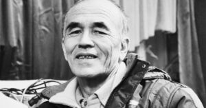 Black and white portrait of Azimjan Askarov, Kyrgyz human rights defender and artist, wearing a jacket and a collared shirt. He is seated indoors, looking toward the camera with a calm, gentle expression.