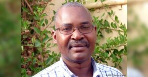 A close-up portrait of Nigerien human rights defender and journalist Moussa Tiangari standing in front of greenery. He is wearing glasses and a white and grey checkered shirt. Credit: DW