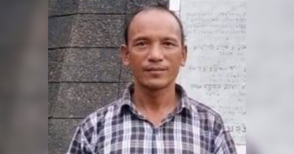 Portrait of environmental human rights defender and indigenous community leader Ringrong Mro, wearing a white and grey checkered shirt and standing in front of a grey wall. He is looking straight into the camera with a faint smile.