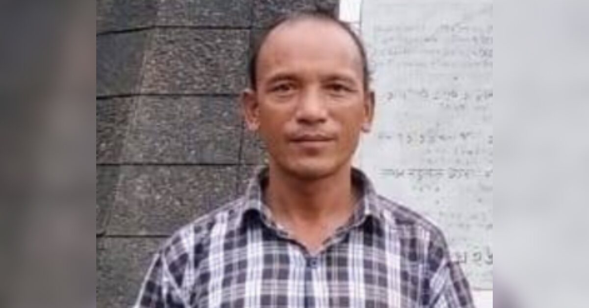 Portrait of environmental human rights defender and indigenous community leader Ringrong Mro, wearing a white and grey checkered shirt and standing in front of a grey wall. He is looking straight into the camera with a faint smile.