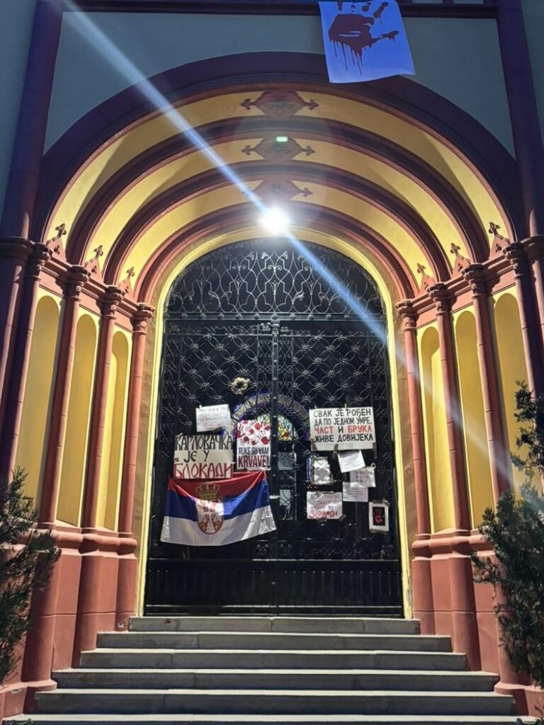 Nighttime view of a high school’s arched entrance in Serbia. The closed wrought-iron gate is covered with protest placards and flyers in Serbian, with a Serbian tricolour flag draped across the centre.