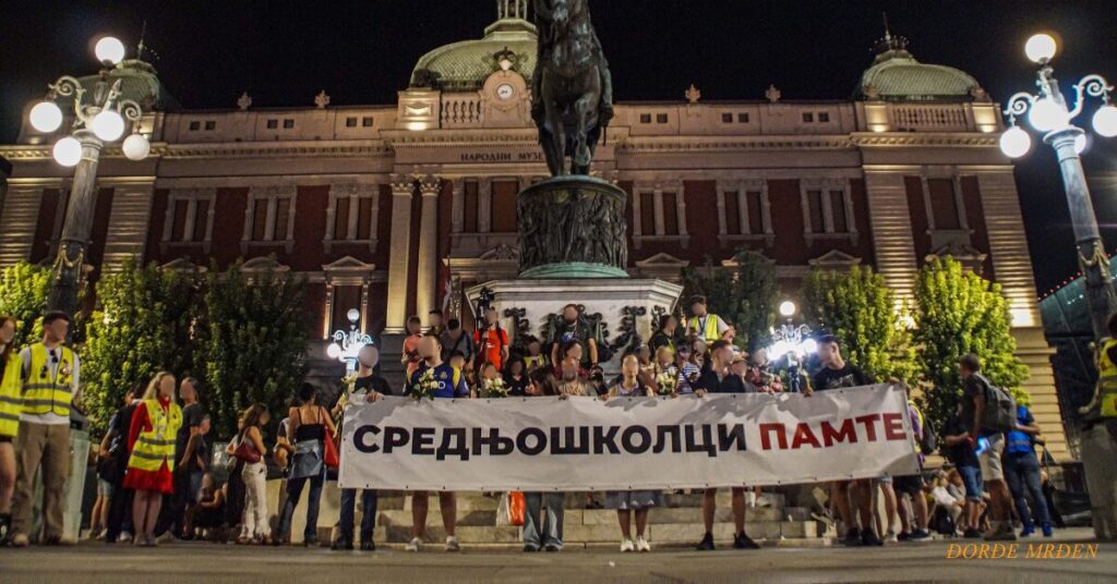 Nighttime protest by Serbian high-school students at Republic Square in Belgrade, in front of the National Museum and the Prince Mihailo equestrian statue. A row of students, some in yellow vests, hold a large white banner in Cyrillic reading “СРЕДЊОШКОЛЦИ ПАМТЕ” (“High-school students remember”), with flowers in hand, while onlookers gather under bright street lamps. Photo by Đorđe Mrđen.