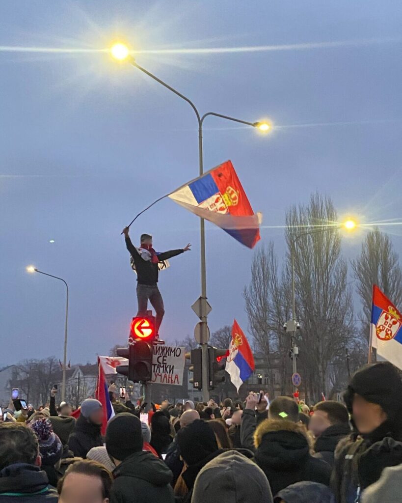 At dusk during a street protest in Serbia, a large crowd in winter coats fills an intersection. A protester stands atop a red traffic light, arms outstretched, waving a Serbian tricolour flag with the coat of arms. More Serbian flags rise from the crowd as people film on phones. Streetlights glow against a grey sky; leafless trees and buildings line the background.
