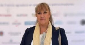 Mexican woman human rights defender Virginia Garay Cazares stands in front of a blurred backdrop with various logos. She has light brown hair with bangs, wears a dark blazer over a light shirt, and a cream-colored scarf around her neck, facing the camera with a smile.
