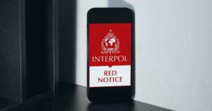A smartphone propped against a white wall displaying the logo of Interpol on a red background, with the mention 'Red notice' in red text over a white background underneath it.
