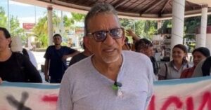 Mexican human rights defender Luis García Villagrán stands outdoors with a group of people holding a large banner. The group is gathered under a shaded pavilion in a public square, with trees and buildings visible in the background. Villagrán is wearing sunglasses and a light grey shirt.