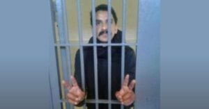 Sahrawi human rights defender Mohamed Embarek Lefkir stands behind metal prison bars, wearing dark clothing. He faces the camera and raises both hands, forming peace signs with his fingers.