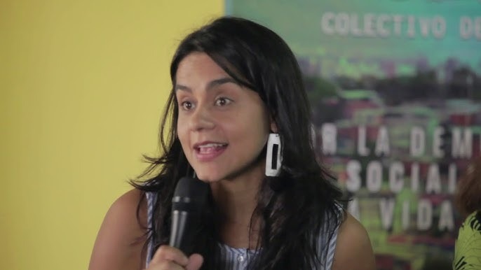 Venezuelan woman human rights defender and lawyer Martha Lía Grajales speaks into a handheld microphone during a public event. She is shown from the shoulders up, wearing a sleeveless light-blue top and large rectangular white earrings, with long dark hair. A yellow wall and a green poster with partially visible Spanish text appear in the background.