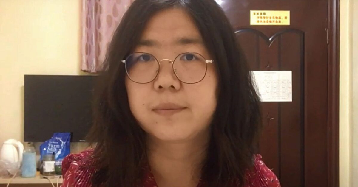 Chinese woman human rights defender Zhang Zhan sits indoors facing the camera, wearing round glasses and a red patterned blouse. She has shoulder-length dark hair and a calm but serious expression. Behind her is a wooden door with a small sign in Chinese, a television screen, and a desk with household items.
