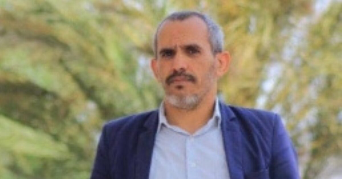 Yemeni human rights lawyer Abdulmajeed Sabra standing outdoors with palm trees in the background. He is wearing a light blue shirt and navy blue suit, and looking straight at the camera with a serious demeanor.