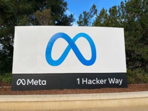 A sign at META headquarters with a blue infinity loop against a white background and below, along a black strip, "Meta", another infinity loop, and the address 1 Hacker Way