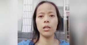 Close-up portrait of Chinese woman human rights defender Yang Li speaking directly to camera outdoors. She appears serious and focused, with dark hair loosely tied back, wearing a blue top. A building entrance and windows are visible behind her, slightly out of focus.