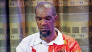 A close up photo of South African human rights defender Zweli “Khabazela” Mkhize, looking down. He is wearing a red and white shirt, and is standing indoors in front of green curtains.